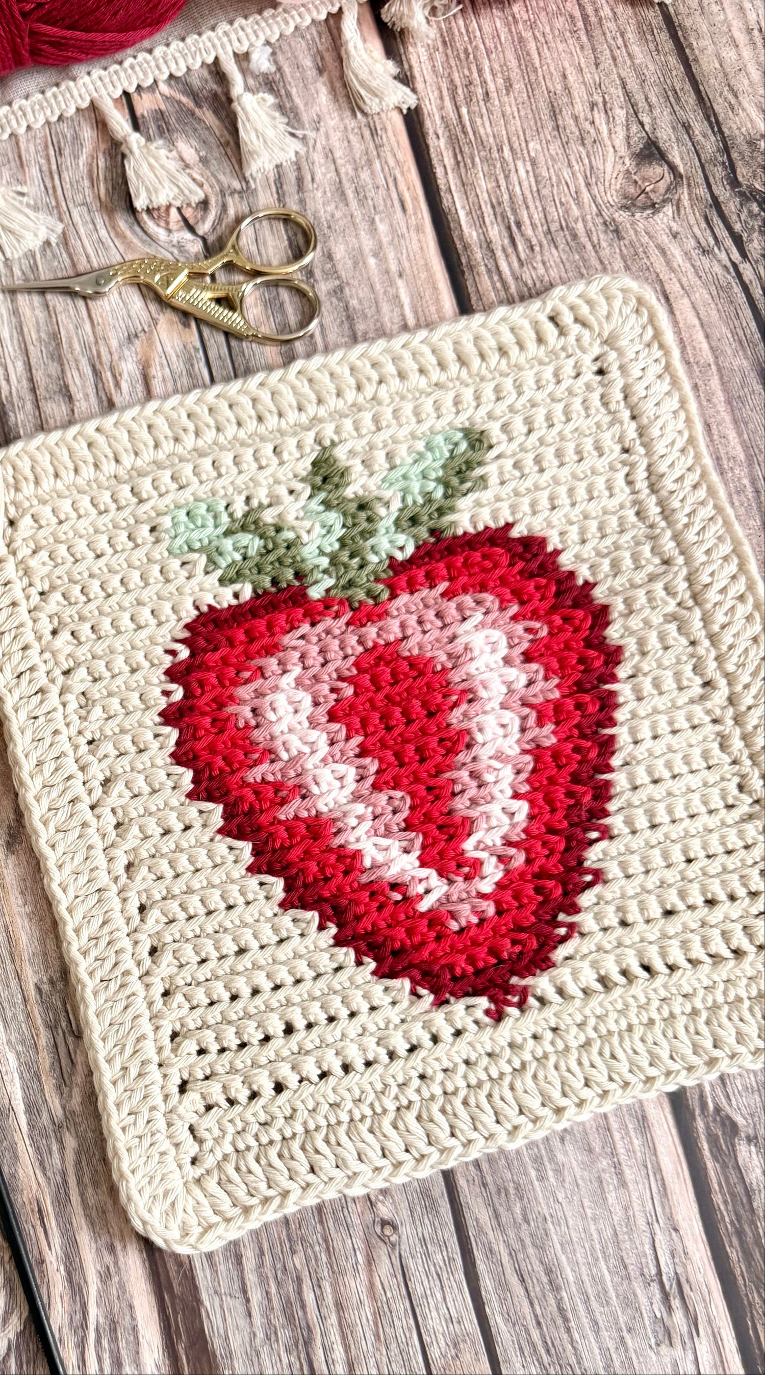 Crocheted strawberry pattern on a wooden surface with yarn and crochet hooks.